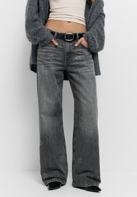 Széles szárú szürke farmernadrág csillag hímzéssel, magas derékkal és övhurkokkal. Bézs felsővel és fekete övvel viselve. Texturált anyag.