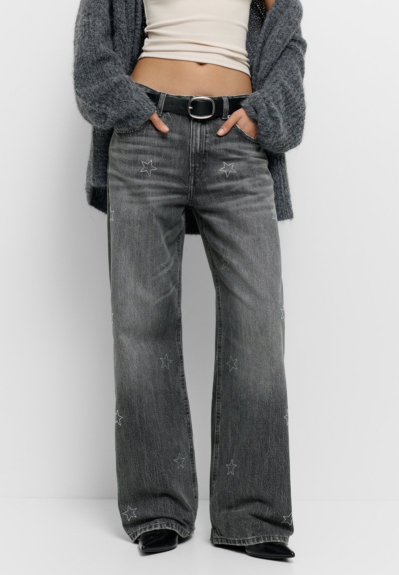 Széles szárú szürke farmernadrág csillag hímzéssel, magas derékkal és övhurkokkal. Bézs felsővel és fekete övvel viselve. Texturált anyag.