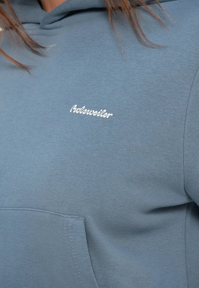Modrá mikina s kapucí a přední kapsou, s bílým kurzívním nápisem "Holzweiler" na hrudi, ukazující část horního trupu a vlasy osoby.