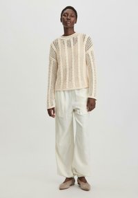 Strickpullover in Creme mit offenen Maschenmustern, langen Ärmeln und rundem Ausschnitt, kombiniert mit lockeren weißen Hosen und beigen Schuhen.