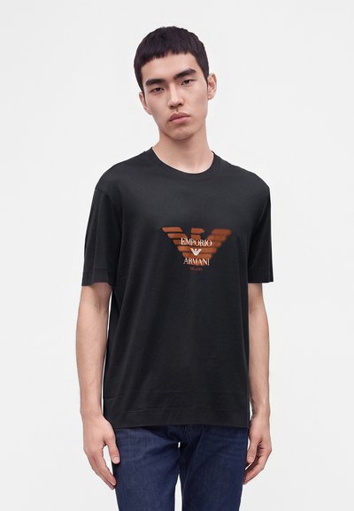 Млад мъж, облечен в черна тениска Emporio Armani с оранжево лого и тъмни дънки, стоящ срещу светъл едноцветен фон.