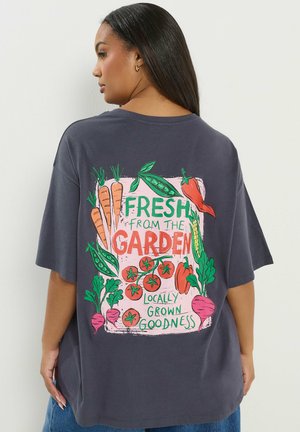 Mujer con cabello largo y oscuro, vestida con una camiseta oscura de gran tamaño con ilustraciones coloridas de verduras y el texto "Fresco del jardín, bondad de cultivo local".
