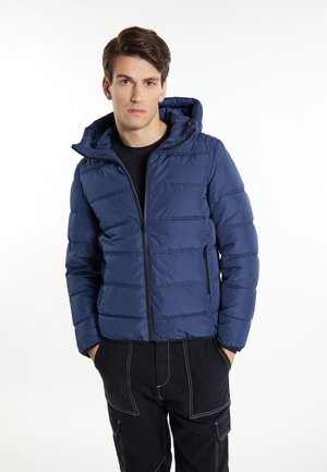 Giovane uomo che indossa una giacca a vento blu navy con cappuccio e pantaloni neri con cuciture bianche, in piedi con le mani nelle tasche su sfondo bianco.