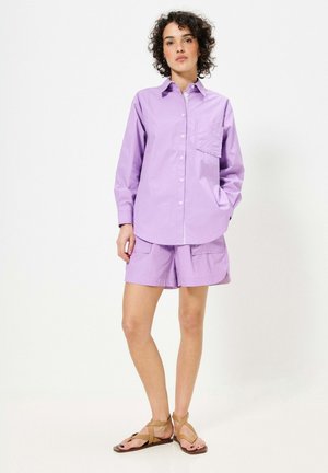 Chemise lavande à boutonnage avec une poche poitrine, associée à un short cargo de style assorti. Des sandales plates achèvent la tenue.