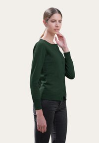 Cardigan vert foncé en matériel doux, avec un col rond, des boutons à l'avant et deux poches latérales. Porté sur un jean foncé.
