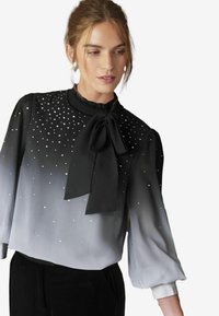 Schwarz- und grauverlaufende Bluse mit langen Ärmeln, verziert mit silbernen Nieten, mit einem rüschierten Ausschnitt und einer großen Schleife an der Vorderseite.