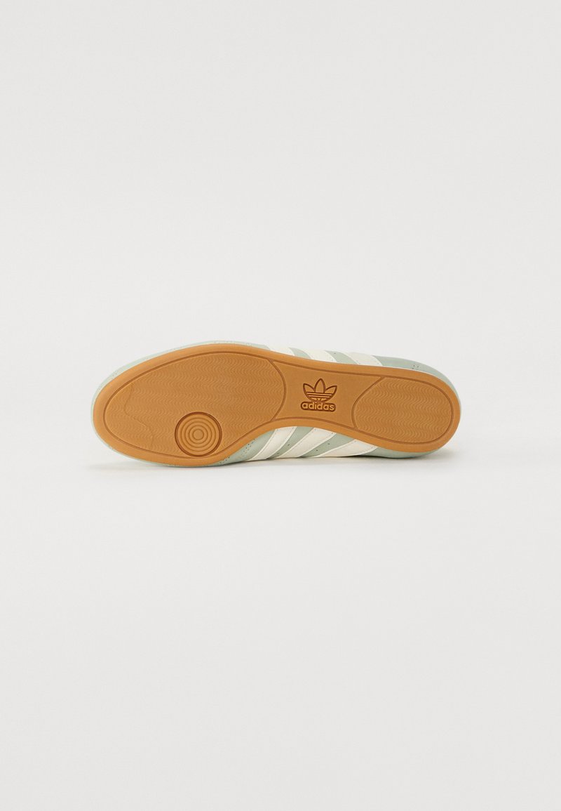 Suela de zapatilla Adidas con rayas en verde claro y crema, superficie de goma texturizada y un logotipo circular marrón. Diseño plano y patrón de tracción moldeado.