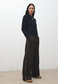 Donna in piedi contro un muro bianco, che indossa un maglione a coste nero con bottoni dorati, pantaloni larghi scuri con strisce laterali e mocassini color sabbia.