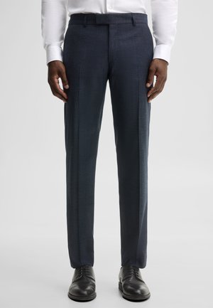 Man draagt donkerblauwe op maat gemaakte pantalon, zwarte nette schoenen en een wit hemd met knopen, staand tegen een effen achtergrond.