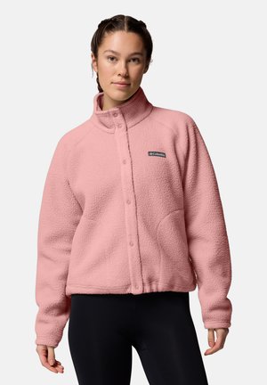 Ung kvinde med flettet hår iført en lyserød Columbia fleecejakke og sorte leggings, stående mod en lys baggrund.
