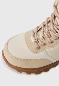 Beige fjellsko med en teksturert kanvas- og lærutside, med beige snøringer akkompagnert av røde og blå tråder og en robust gummisåle.