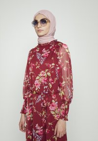 Blusa floral fluída em bordô com mangas longas e transparentes e punhos franzidos; combinada com um hijab rosa claro e óculos de sol oversized acentuados com dourado.