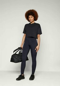 T-shirt cropped nero e leggings abbinati, entrambi realizzati in materiale elastico. La modella tiene in mano una borsa a tracolla nera. Le scarpe da ginnastica sono nere.