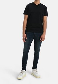 Svart v-ringad t-shirt i bomull, kombinerad med mörkblå jeans. Modellen bär vita sneakers med gula sulor och en slät yta.