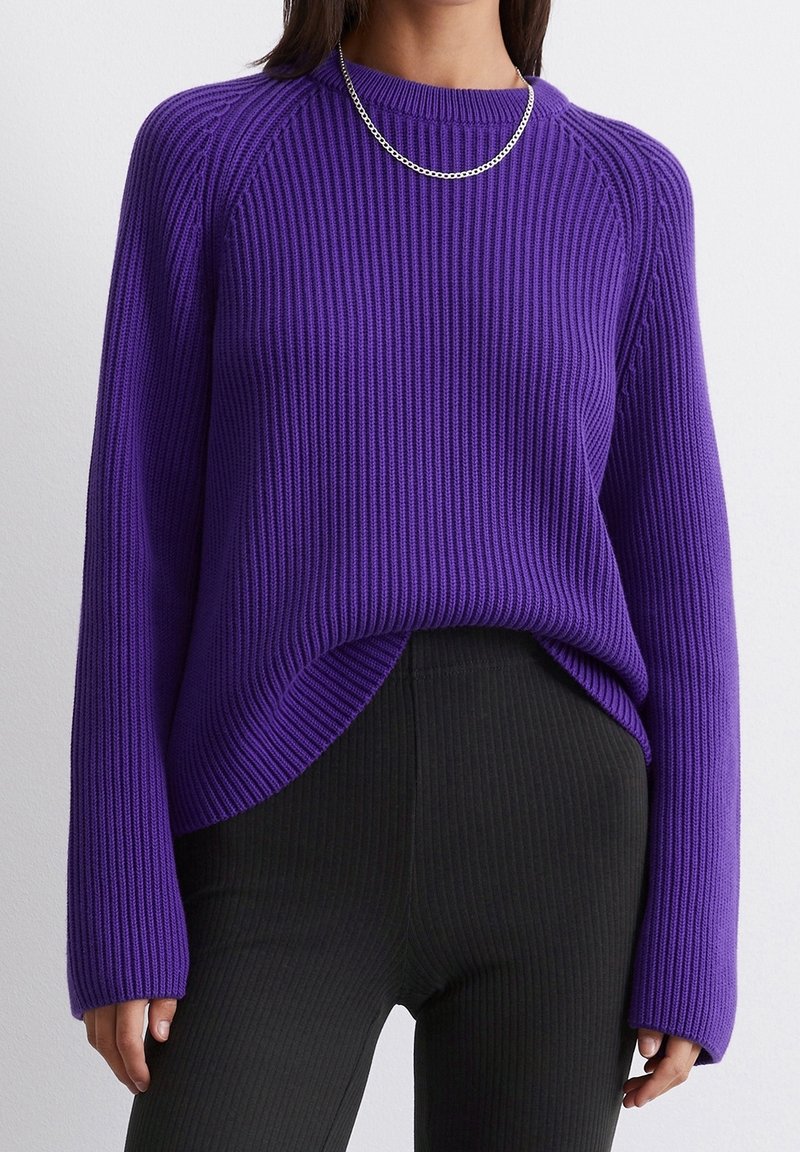 Femme portant un pull violet côtelé à manches longues, un pantalon noir côtelé et un collier chaîne argenté sur un fond uni.