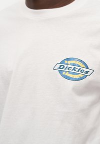 Λευκό βαμβακερό t-shirt με μπλε και κίτρινο κυκλικό σχέδιο και το κείμενο "Dickies" στο κέντρο. Κοντά μανίκια και στρογγυλή λαιμόκοψη.