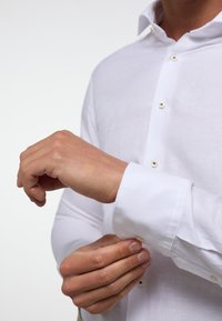 Witte overhemd met knoopsluiting en kraag, gladde textuur en lange mouwen. Manchetjes vastgezet met knopen terwijl de hand de mouw aanpast.