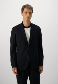 Mand med kort, blond hår iført en sort blazer over en hvid ribbet skjorte, der står mod en ensfarvet lys baggrund.