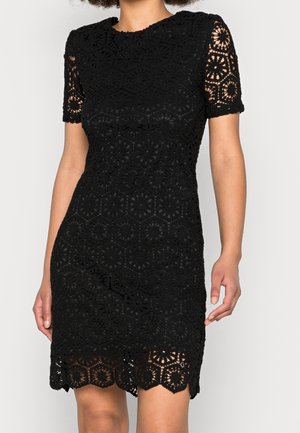 Vrouw draagt een nauwsluitende zwarte kanten jurk met korte mouwen en een geschulpte zoom, met een geometrisch bloemenpatroon over de hele jurk.