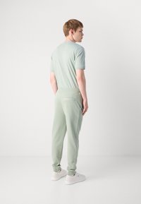 Joven de pie de lado, vistiendo una camiseta casual verde clara, pantalones jogger a juego y zapatillas blancas, frente a un fondo liso.