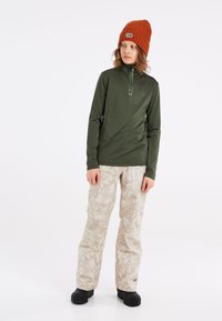 Camiseta verde de manga larga con cremallera y logotipo, combinada con pantalones estampados color blanco roto. El niño lleva un gorro naranja texturizado y botas negras.