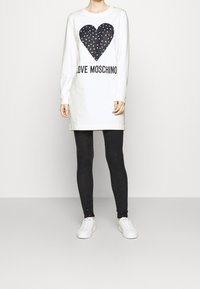 Bílé šaty s dlouhým rukávem a černým srdcem na potisku, s barevnými akcenty a textem "LOVE MOSCHINO". Spárováno s černými strukturovanými legínami a bílými teniskami.