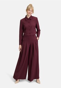 Completo due pezzi bordeaux composto da un giubbotto corto con colletto tondo e grandi bottoni, abbinato a pantaloni a gamba larga con pieghe realizzati in tessuto morbido.