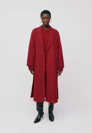 Red long coat with a structured silhouette, wide lapels, and side pockets, paired with a textured red sweater and knee-high black boots.