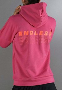 Persona con una sudadera rosa que tiene la palabra "ENDLESS" en letras gruesas y contorneadas en la parte posterior, ajustándose la capucha.