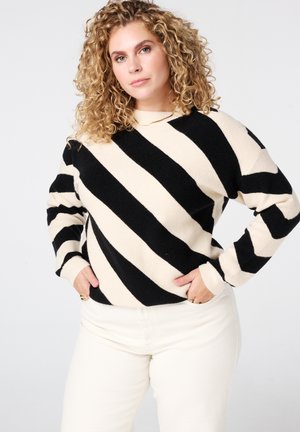 Vrouw met krullend haar, gekleed in een zwart-wit diagonale gestreepte trui en lichtgekleurde broek, staand met handen in de zakken tegen een witte achtergrond.