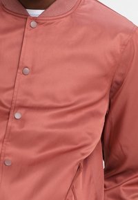 Close-up of a person wearing a dusty rose satin bomber jacket with snap buttons and a ribbed collar over a white shirt.
