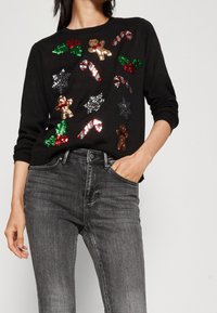 Schwarzer Pullover mit Pailletten in Form von Lebkuchenmännern, Zuckerstangen, Stechpalme und Schneeflocken in verschiedenen Farben. Kombiniert mit grauen Jeans.