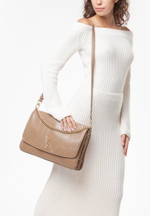 Mujer con vestido de punto blanco sin hombros, sosteniendo un bolso de hombro de cuero topo con cadena dorada y emblema con logo.