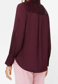 Bordeauxrød bluse med lange ærmer og en glat tekstur, der har et bærestykke, plissering bagpå og manchetter med knapper.