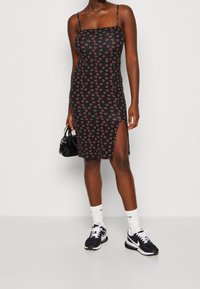 Schwarzes ärmelloses Minikleid mit Kirschmuster, seitlichem Schlitz und tailliertem Schnitt; kombiniert mit schwarzen Sneakers und weißen Socken.