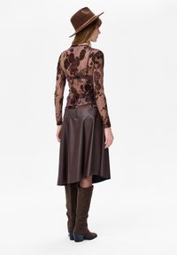 Vrouw die een bruin bloemen doorschijnend topje draagt, een donderbruine leren rok, kniehoge bruine suède laarzen en een bijpassende bruine fedora hoed, staand opzij.