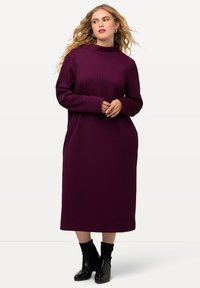 Langärmliges Kleid mit Stehkragen in Dunkelpurpur mit strukturiertem Muster. Hat eine lockere Passform, Seitentaschen und reicht bis unter das Knie, kombiniert mit schwarzen Stiefeletten.