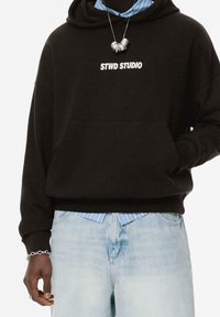 Sweat à capuche noir avec le texte "STWD STUDIO" porté sur une chemise rayée bleue, jean délavé clair, bracelet chaîne en argent et collier avec des perles métalliques.