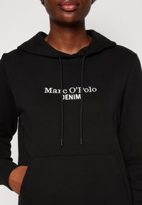 Černá mikina s kapucí, se přední kapsou, stahovacími šňůrkami a bílým vyšitým nápisem "Marc O'Polo DENIM". Vyrobena z měkké látky.