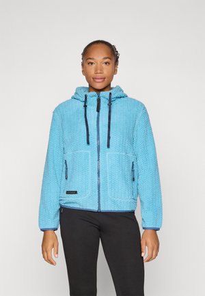 Nő világoskék, texturált cipzáras kapucnis pulóvert visel fekete húzózsinórokkal és fekete nadrágot, előre néz egy egyszerű háttér előtt.