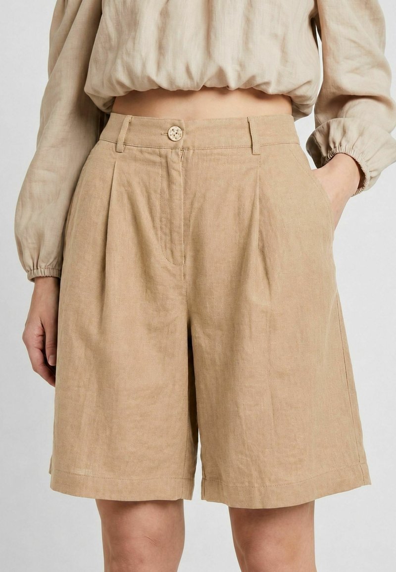 Short en lin beige taille haute avec plis devant, fermeture bouton et zip, poches latérales, porté avec un haut beige à manches longues ample.