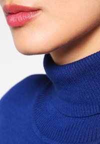Prenda de cuello alto tejida en azul con textura acanalada y diseño ajustado que destaca la zona del cuello. Piel suave visible debajo.