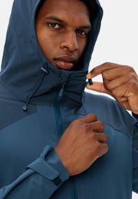 Giacca a vento blu navy con chiusura a zip e cordini regolabili; presenta un tessuto testurizzato e polsini in velcro.