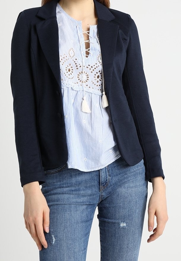 Blazer navy su una blusa a righe azzurra chiara con dettagli ricamati e nappine bianche. Indossato con jeans blu strappati.