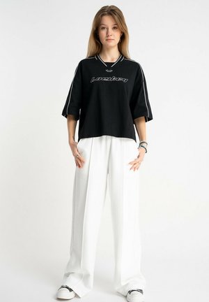 Młoda kobieta z długimi włosami, ubrana w czarną, oversize'ową koszulę z białymi napisami, białe spodnie o szerokich nogawkach oraz białe trampki, stojąca na tle białego tła.