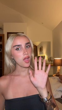 Jeune femme blonde avec de longs cheveux, portant un haut sans bretelles noir et un bracelet en argent, levant la main dans une chambre moderne bien éclairée.