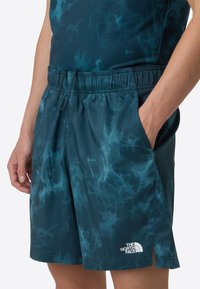 Swim shorts in dark teal with a water-like pattern, elastic waistband, drawstring, and side pockets. Features a logo on the hem.