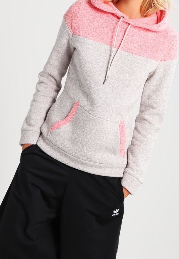 Personne portant un sweat à capuche gris clair et rose avec poches avant et un pantalon noir Adidas avec un logo sur la cuisse gauche.