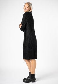 Schwarz geripptes, knielanges Strickkleid mit hohem Kragen und langen Ärmeln, kombiniert mit klobigen, schwarzen Ankle Boots mit Absatz.