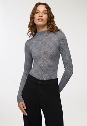 Giovane donna con capelli ricci che indossa un dolcevita grigio fitted con motivo e pantaloni neri a vita alta su uno sfondo semplice.